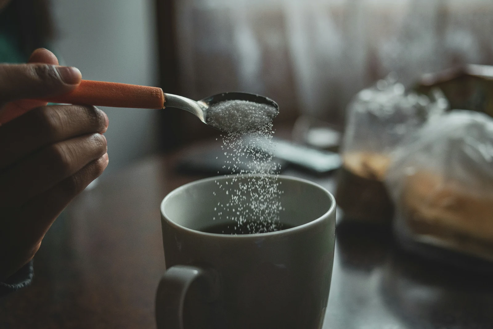 O mână înclină o linguriță de zahăr deasupra unei cești de cafea, zahărul căzând în șuviță spre suprafață