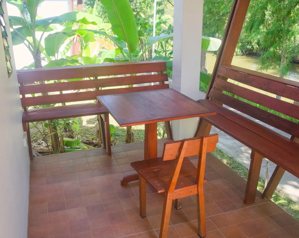 Veranda casei din Thailanda, cu masă și bănci de lemn, la umbră, cu bananieri și un canal în fundal